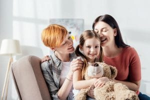 Smiling kid with teddy bear looking at camera near happy same sex parents on sofa at home