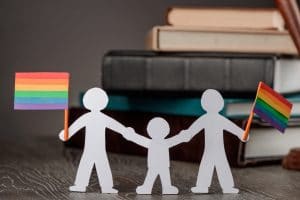 Paper cutter of same-sex couple holding hands with child and holding pide flags with stacked book background.