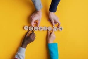 Hands of a family with black and white children assembling the word "Adoption" spelled on wooden cut circles over yellow background.