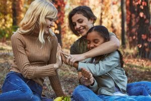 A beautiful couple of lesbian ladies having fun in the park with their adopted teenage daughter. The young family sitting on the ground, playing patty-cake, laughing. Autumn trees in the background.