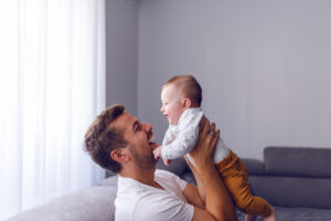 Playful handsome caucasian young dad lifting his loving 6 months old baby boy while sitting on sofa in living room. Baby enjoying time with father and laughing.