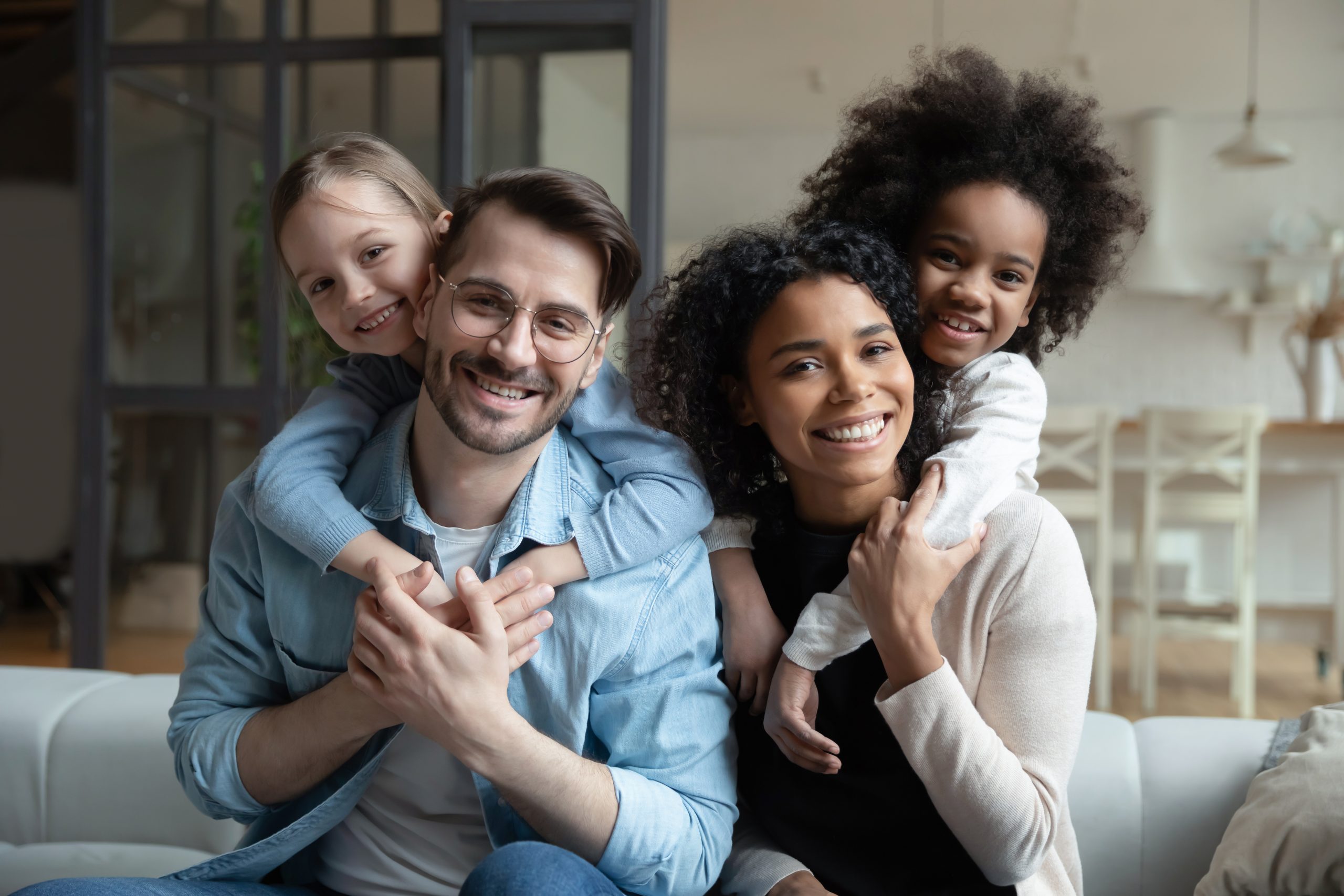 Portrait of happy young multiethnic family with small diverse daughters relax in living room at home. Smiling multiracial mom and dad cuddle hug with adopted small girls children. Adoption concept.