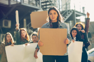 Young woman with blank poster in front of people protesting about women's rights and equality on the street. Meeting about problem in workplace, male pressure, domestic abuse, harassment. Copyspace.