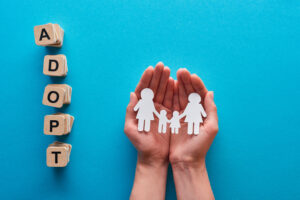 cropped view of woman holding paper cut lesbian family near blocks with adopt lettering on blue background