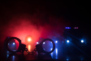 Police raid at night and you are under arrest concept. Silhouette of handcuffs with police car on backside. Image with the flashing red and blue police lights at foggy background. Slider shot